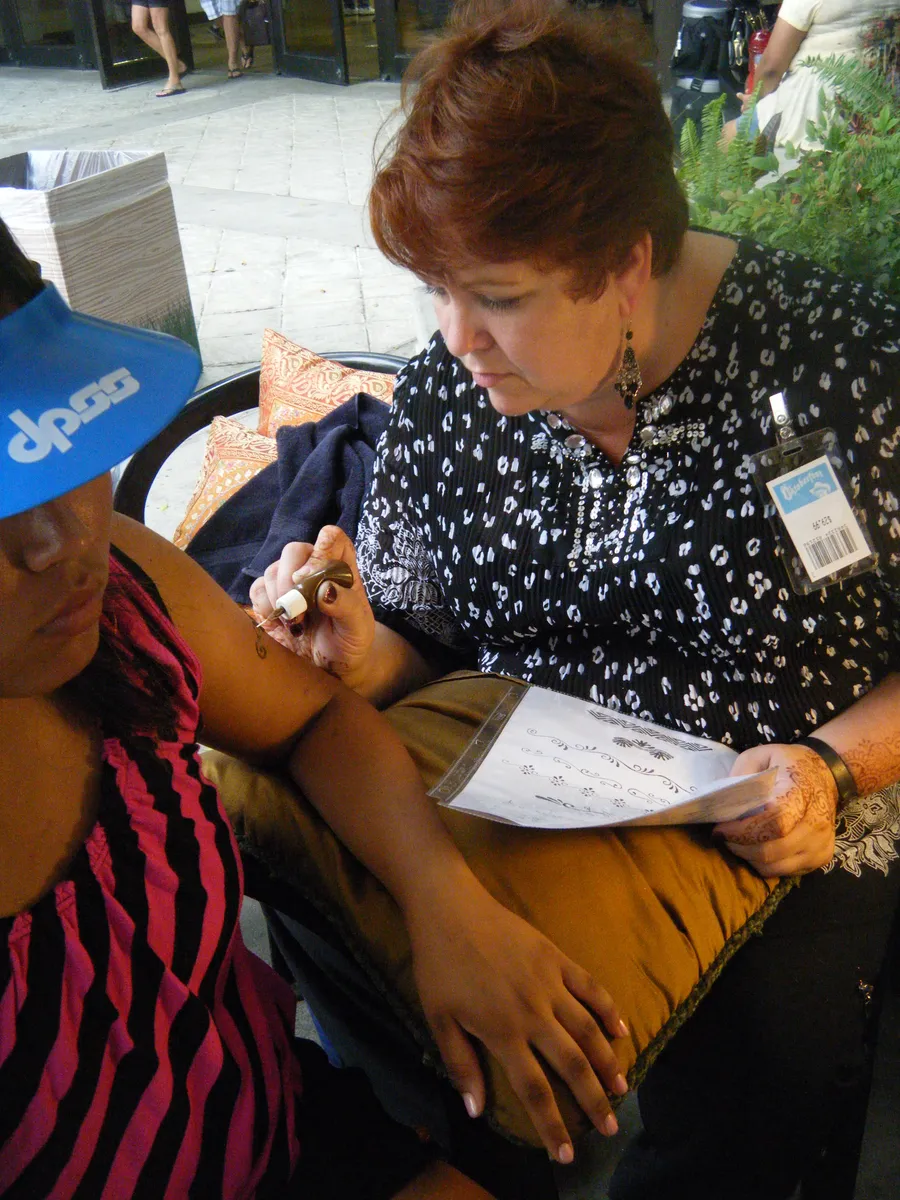 Cynthia MacConnell applying a henna design to a guest's arm at an outdoor event, her design book resting on her lap