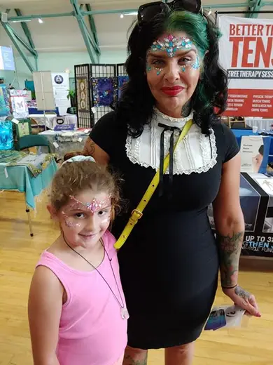 A young guest and an adult guest both wearing gem-and-glitter glam designs at a fair