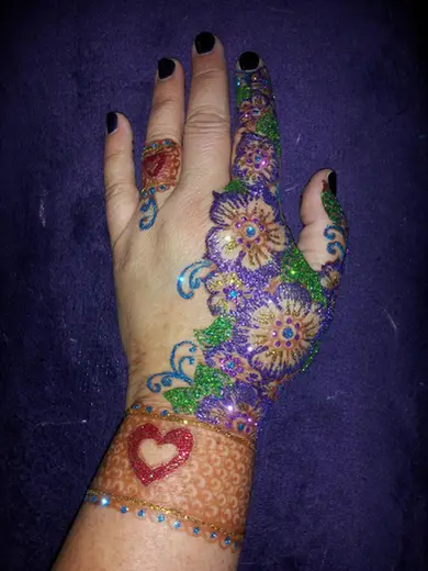 A hand covered in freehand glitter flowers over a henna cuff, with a red glitter heart at the wrist