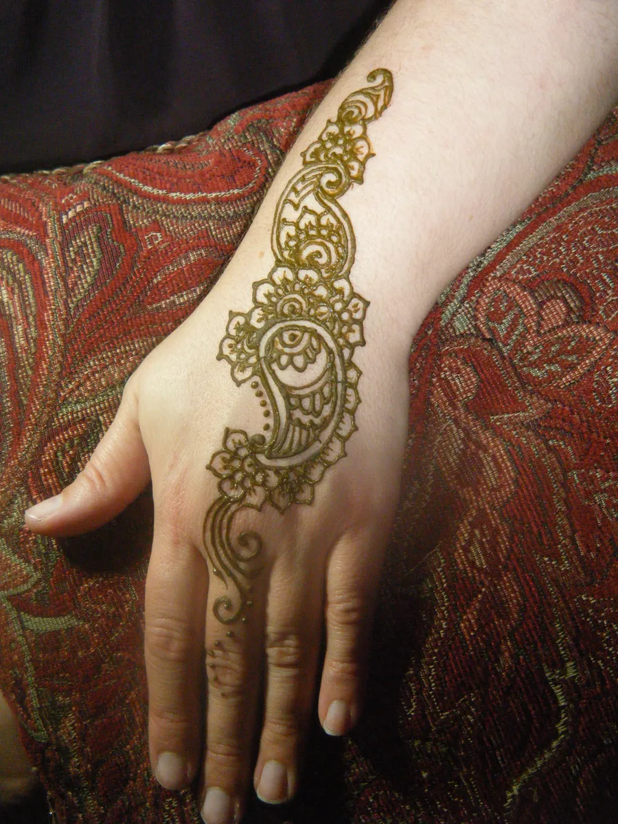 A hand decorated with a fresh green henna paisley and floral design, resting on a red patterned cushion