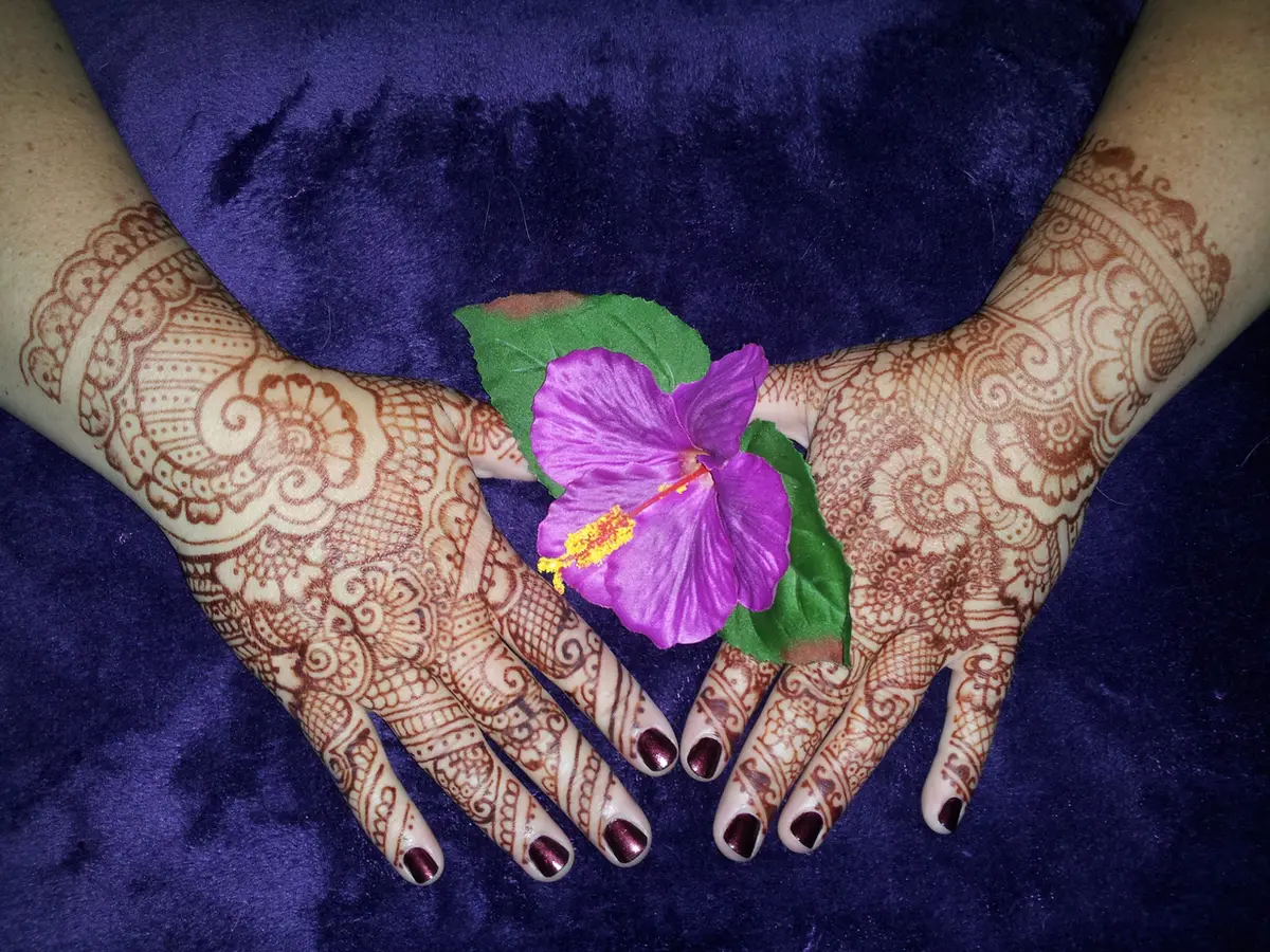 Two hands covered in intricate reddish-brown henna designs resting on purple velvet beside a purple hibiscus flower